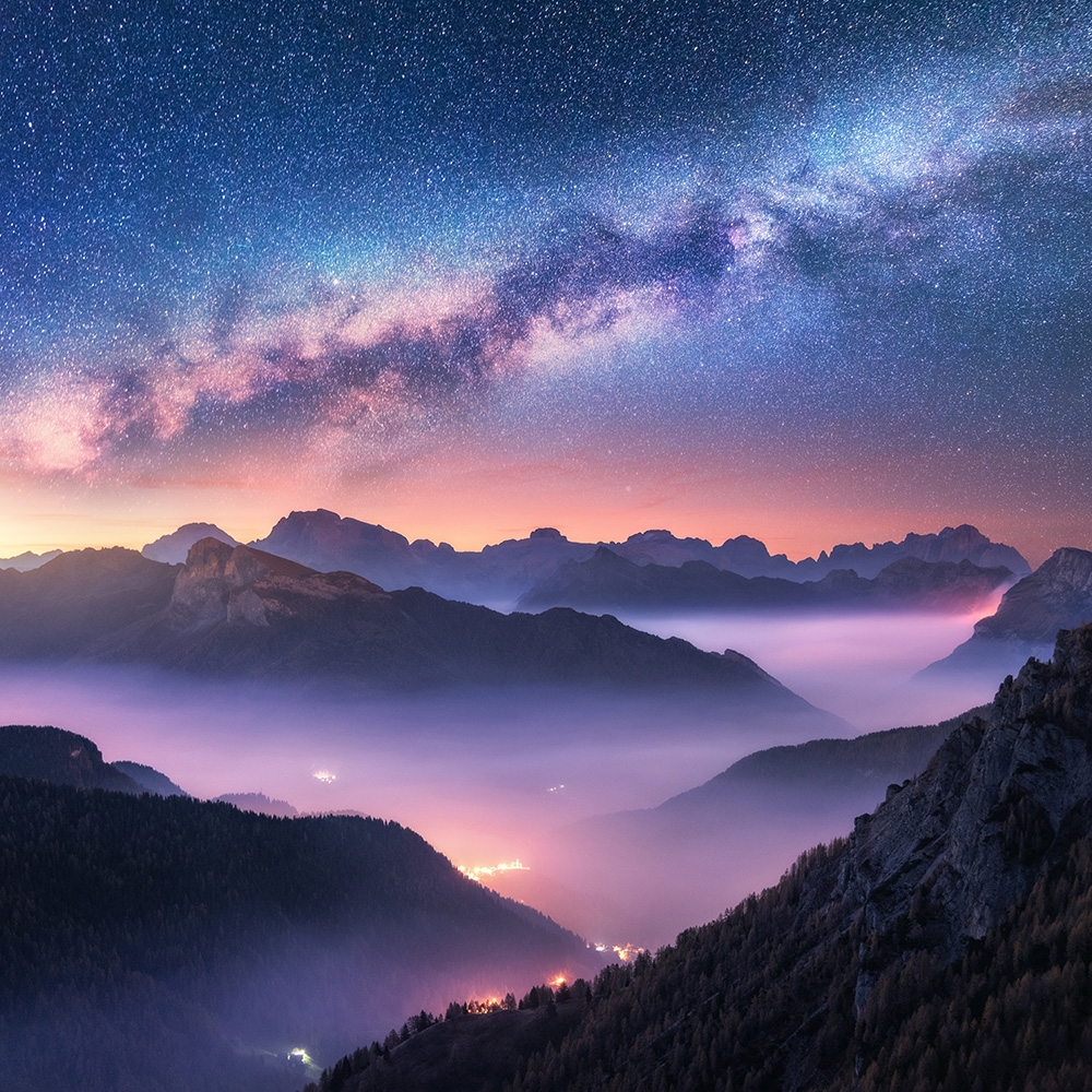 Papier peint photo - Vue à couper le souffle d'un paysage de montagne sous une voie lactée scintillante la nuit, avec des vallées illuminées dans le brouillard