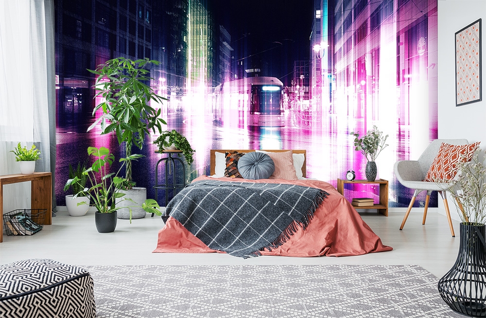 Papier peint photo - Chambre à coucher stylée, décorée avec des plantes et un lit devant un papier peint photo avec un motif urbain de nuit en rose et violet.