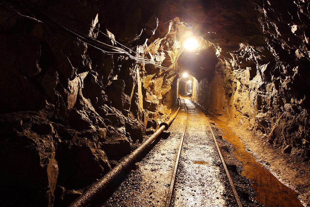 Papier peint photo - Une galerie de mine éclairée avec des rails passe par un tunnel rocheux, des murs de roche rugueuse entourent le chemin.
